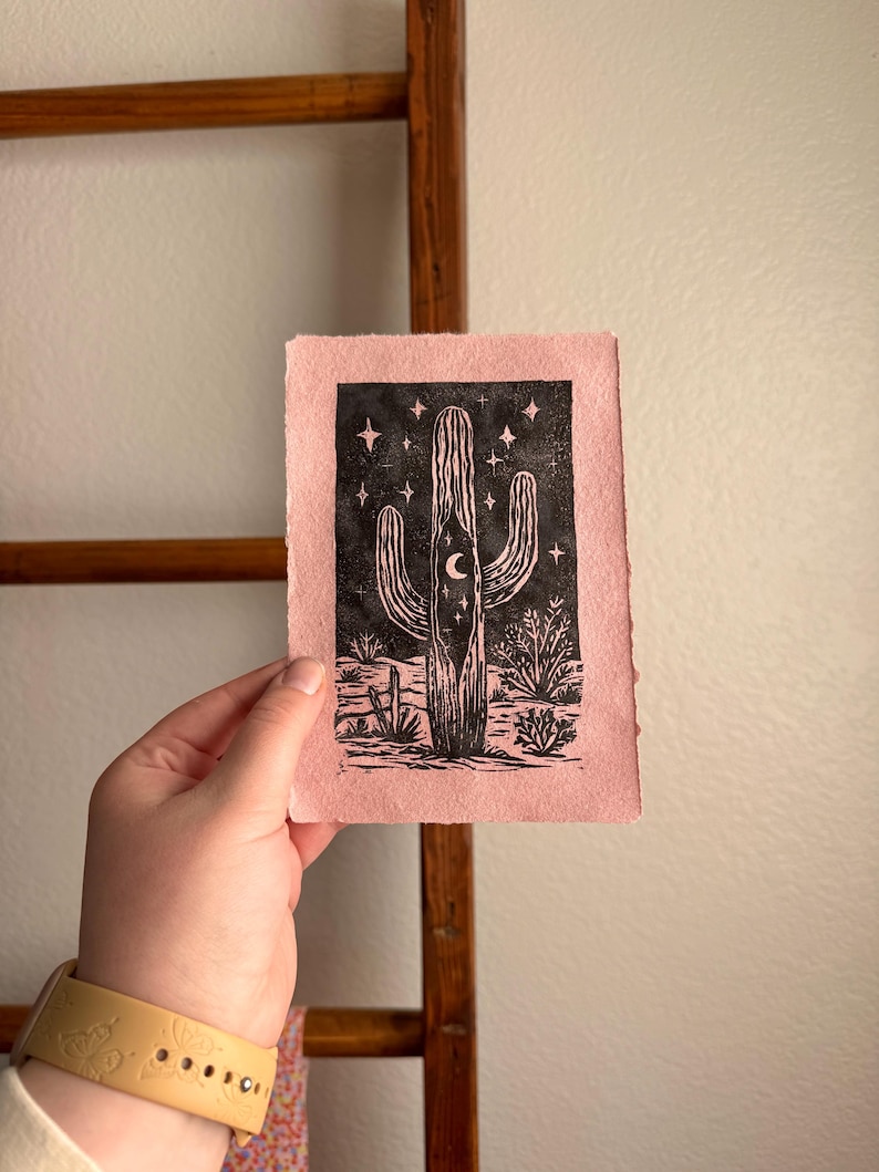 May include: A pink print featuring a black cactus design with a crescent moon and stars. The artwork is a linocut print, showcasing a desert landscape. The print is held against a wooden ladder.
