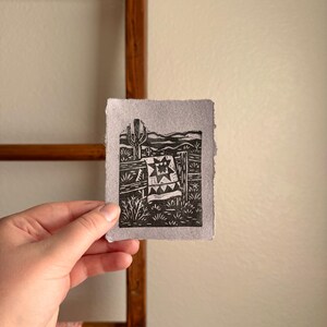 May include: A black and white linocut print depicting a desert scene with a cactus, fence, and a quilt. The print is held in a hand, with a wooden ladder and a patterned fabric in the background.