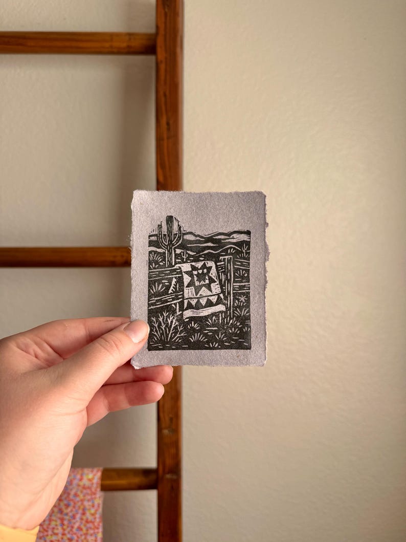 May include: A black and white linocut print depicting a desert scene with a cactus, fence, and a quilt. The print is held in a hand, with a wooden ladder and a patterned fabric in the background.