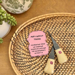 May include: Two wood-handled linocut tools with silver blades rest on a woven wicker tray. A pink card with the text "MAY LINOCUT TEASER" and other text is also on the tray. A decorative white and green vase is in the background.