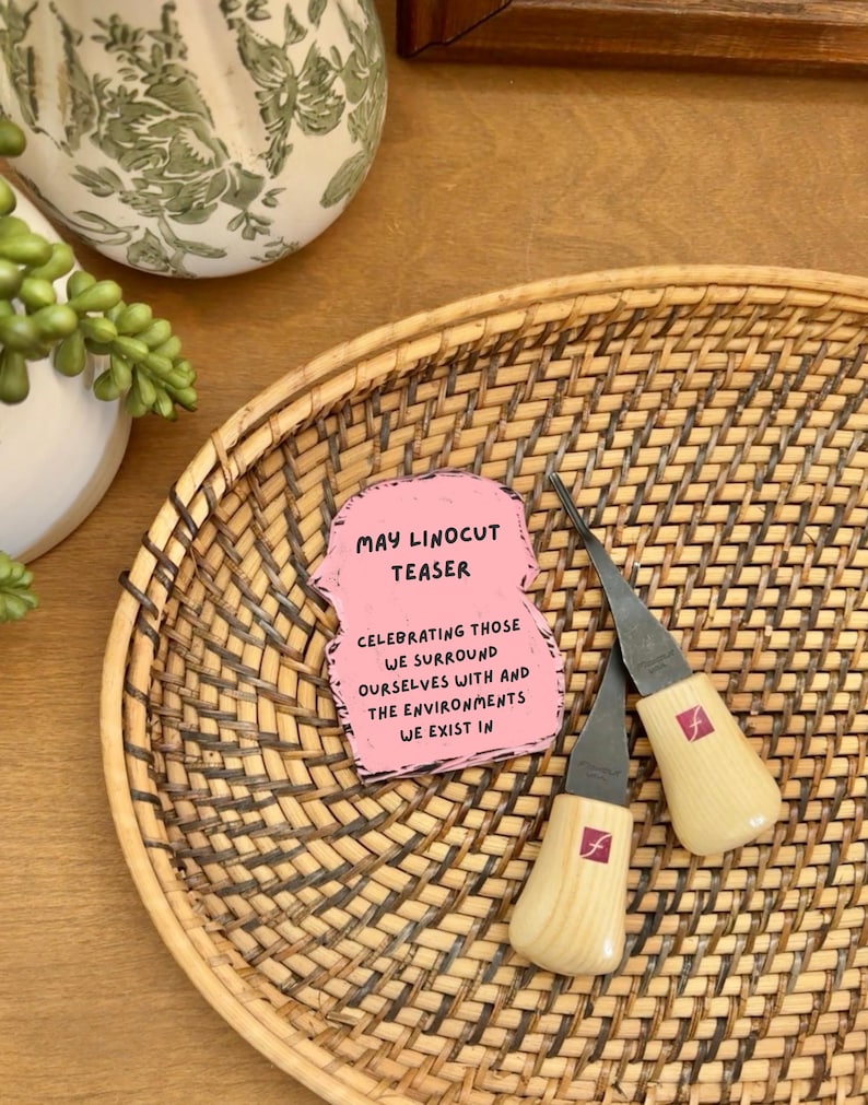 May include: Two wood-handled linocut tools with silver blades rest on a woven wicker tray. A pink card with the text "MAY LINOCUT TEASER" and other text is also on the tray. A decorative white and green vase is in the background.