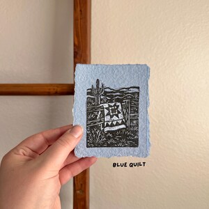 May include: A small, light blue print with a black and white linocut design. The artwork depicts a desert scene with a cactus, fence, and a quilt. The text "BLUE QUILT" is printed in black at the bottom of the print.
