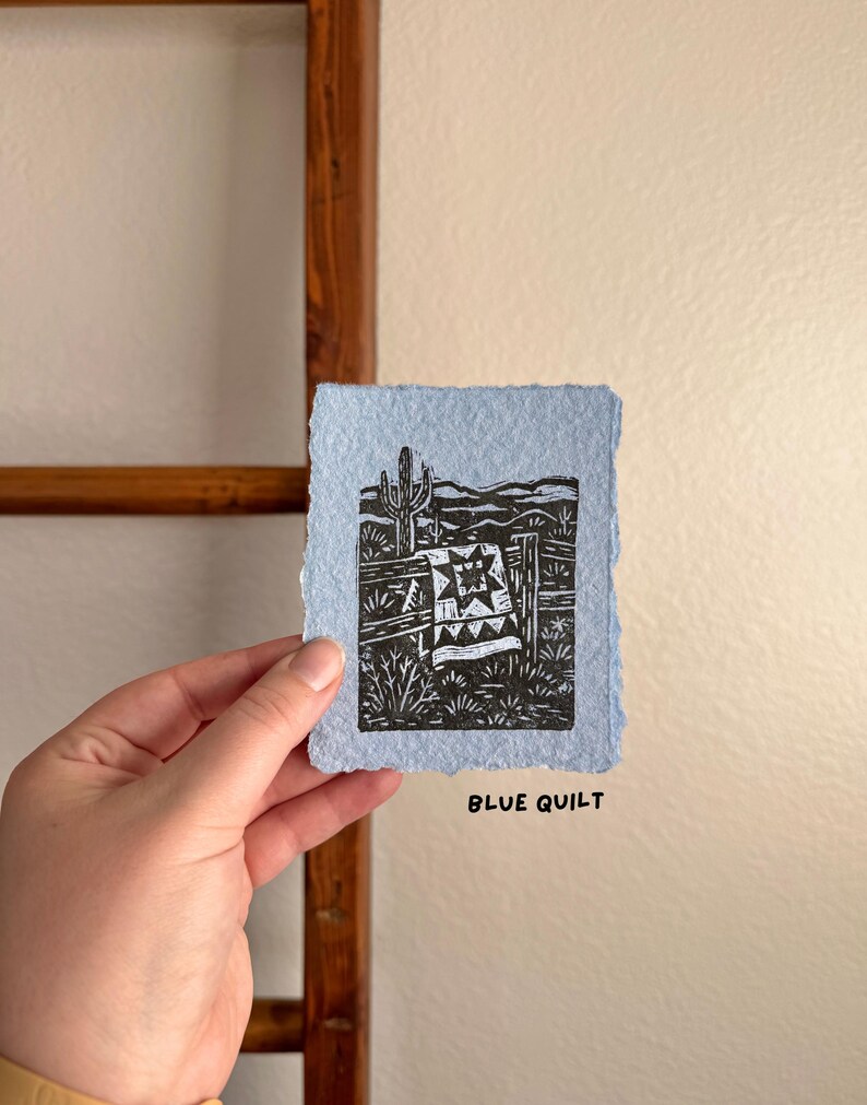 May include: A small, light blue print with a black and white linocut design. The artwork depicts a desert scene with a cactus, fence, and a quilt. The text "BLUE QUILT" is printed in black at the bottom of the print.