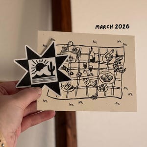May include: A black and white star-shaped sticker with a desert landscape design is held next to a beige card with a picnic illustration. The card includes the text "MARCH 2026". The picnic scene features food, drinks, and a lizard.