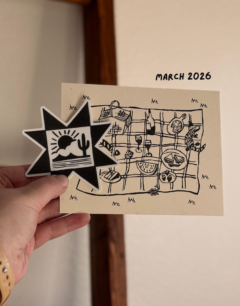 May include: A black and white star-shaped sticker with a desert landscape design is held next to a beige card with a picnic illustration. The card includes the text "MARCH 2026". The picnic scene features food, drinks, and a lizard.