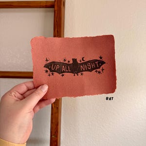 May include: A hand-held, rectangular, reddish-orange paper print with a black bat illustration and the words "UP ALL NIGHT". The bat is surrounded by stars and a crescent moon. The word "BAT" is printed in black.