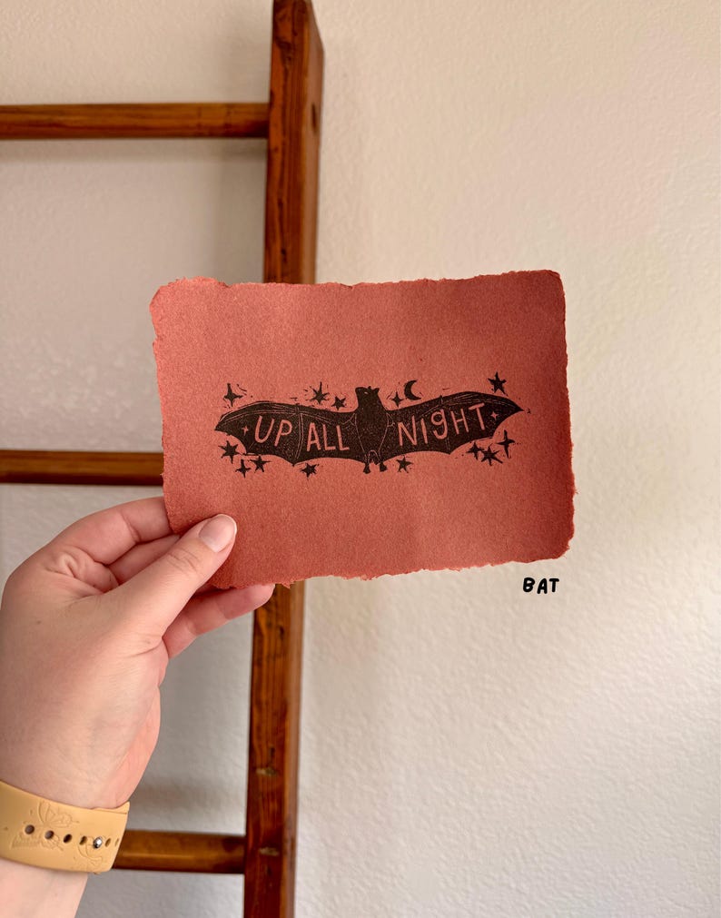 May include: A hand-held, rectangular, reddish-orange paper print with a black bat illustration and the words "UP ALL NIGHT". The bat is surrounded by stars and a crescent moon. The word "BAT" is printed in black.