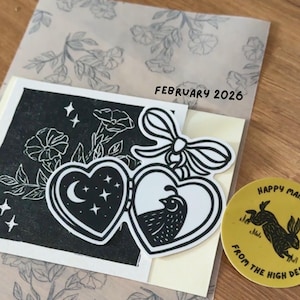 May include: A black and white illustration featuring two heart-shaped designs with a moon and stars, and a bird. The illustration is on a cream-colored card, with the text "FEBRUARY 2026". A yellow sticker with a rabbit is also visible.