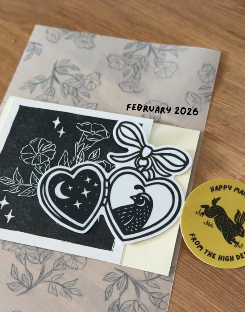 May include: A black and white illustration featuring two heart-shaped designs with a moon and stars, and a bird. The illustration is on a cream-colored card, with the text "FEBRUARY 2026". A yellow sticker with a rabbit is also visible.