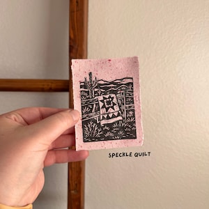 May include: A small, pink speckled paper print with a black linocut design. The design features a desert landscape with a quilt, cactus, and fence. The text "SPECKLE QUILT" is printed in black below the image.