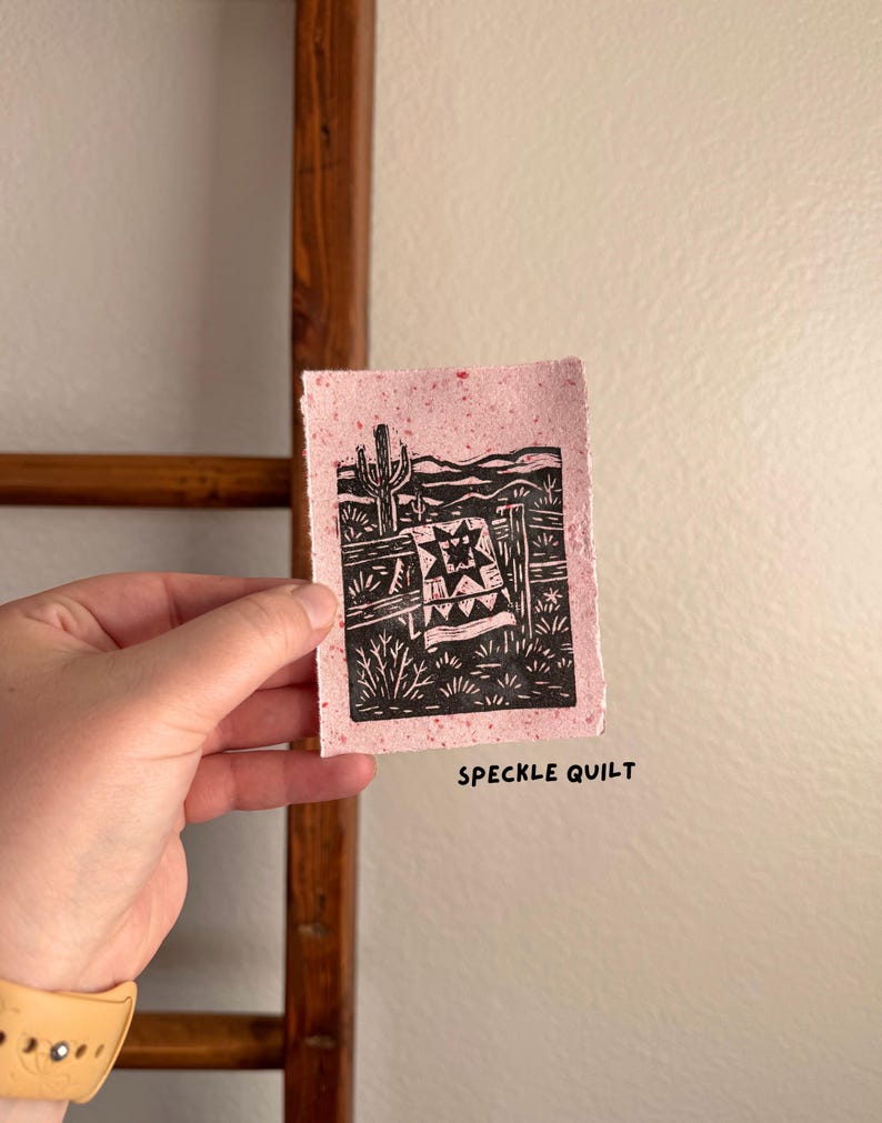 May include: A small, pink speckled paper print with a black linocut design. The design features a desert landscape with a quilt, cactus, and fence. The text "SPECKLE QUILT" is printed in black below the image.