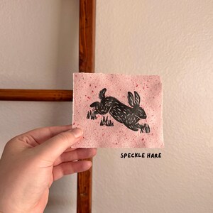 May include: A black linocut print of a running hare on pink speckled paper. The artwork is held in a hand and features the text "SPECKLE HARE" in a simple font. The background is a neutral wall.