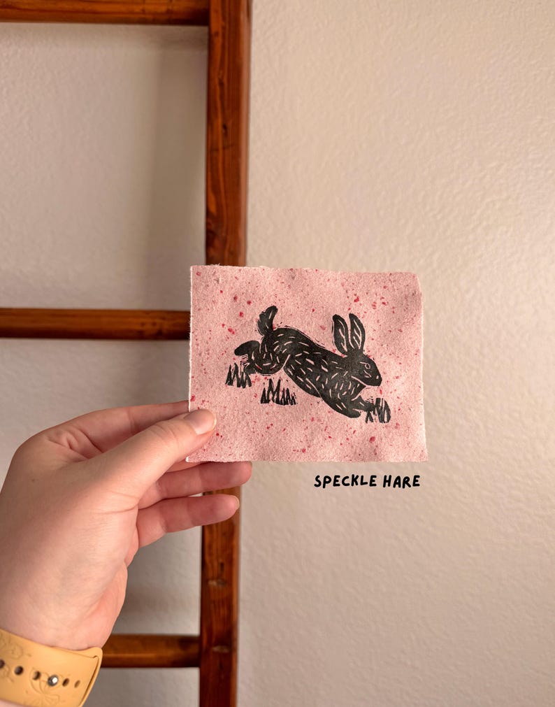 May include: A black linocut print of a running hare on pink speckled paper. The artwork is held in a hand and features the text "SPECKLE HARE" in a simple font. The background is a neutral wall.