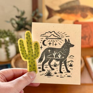 May include: A hand holds a yellow cactus sticker next to a tan card with a black illustration of a coyote, desert landscape, and celestial elements. The artwork has a linocut style.