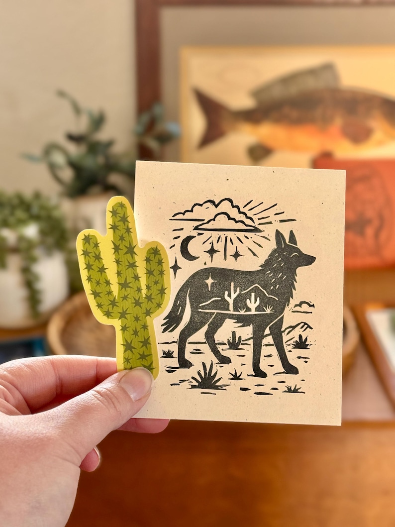 May include: A hand holds a yellow cactus sticker next to a tan card with a black illustration of a coyote, desert landscape, and celestial elements. The artwork has a linocut style.