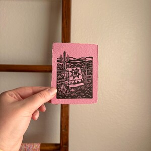 May include: A small, pink print featuring a black and white desert landscape with a cactus, fence, and blanket design. The artwork is held in a hand, showcasing the textured paper and the detailed linework of the print.