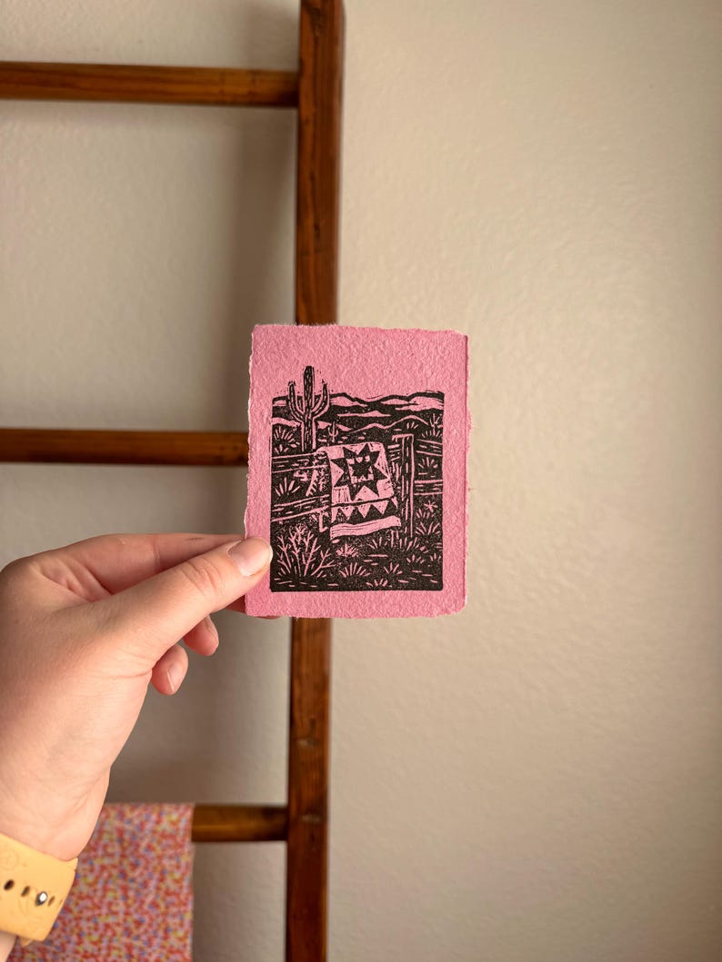 May include: A small, pink print featuring a black and white desert landscape with a cactus, fence, and blanket design. The artwork is held in a hand, showcasing the textured paper and the detailed linework of the print.