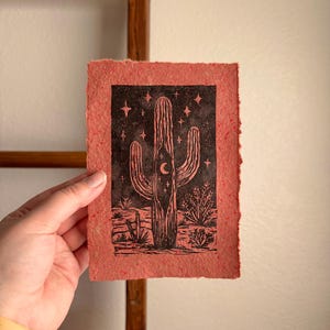 May include: A red-orange print featuring a black illustration of a saguaro cactus with a crescent moon and stars. The cactus is set against a desert landscape. The print has a textured, handmade paper appearance.