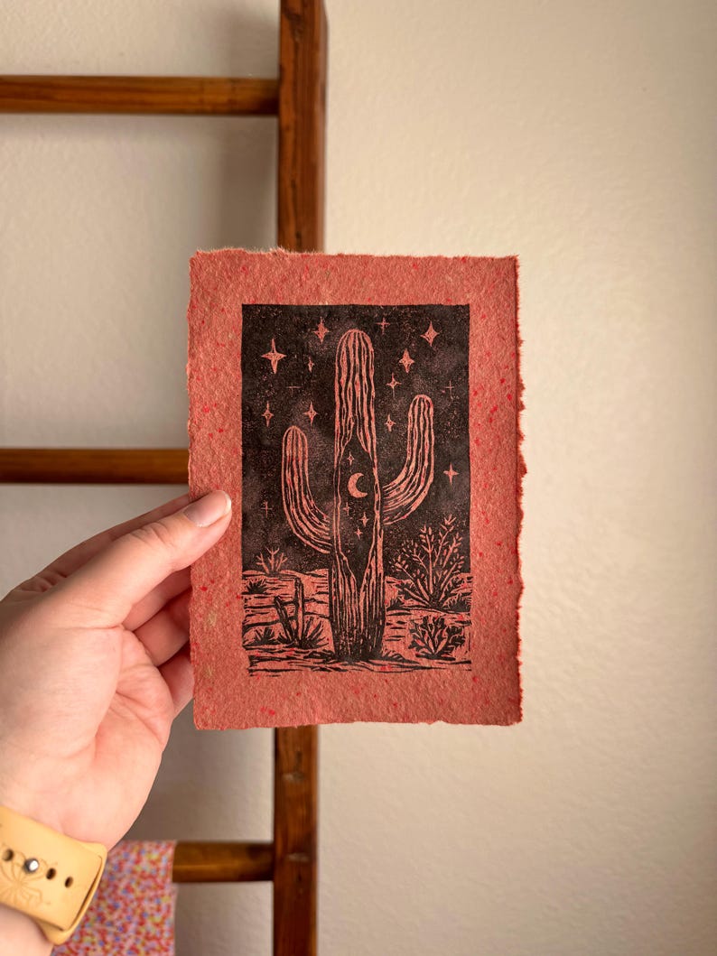 May include: A red-orange print featuring a black illustration of a saguaro cactus with a crescent moon and stars. The cactus is set against a desert landscape. The print has a textured, handmade paper appearance.