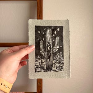 May include: Black and white linocut print of a saguaro cactus under a starry night sky. The print features a crescent moon and desert plants. The artwork is on textured, off-white paper.
