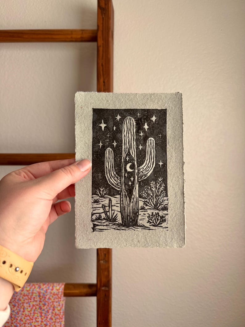 May include: Black and white linocut print of a saguaro cactus under a starry night sky. The print features a crescent moon and desert plants. The artwork is on textured, off-white paper.