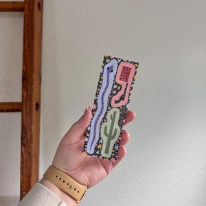 Op de afbeelding: Een rechthoekige boekenlegger met een zwarte achtergrond en een patroon van kleurrijke sterren. Het ontwerp bevat een lange lichtpaarse slang, een roze vorm met een zwart revolver en de tekst HOWDY, en een lichtgroene cactusillustratie.