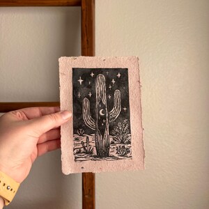 May include: A black and white linocut print of a saguaro cactus with a crescent moon and stars on a pinkish-beige paper. The print depicts desert plants and a fence at the base of the cactus. The paper has a rough, textured appearance.