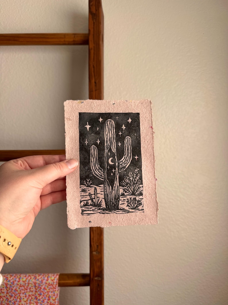 May include: A black and white linocut print of a saguaro cactus with a crescent moon and stars on a pinkish-beige paper. The print depicts desert plants and a fence at the base of the cactus. The paper has a rough, textured appearance.