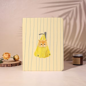 May include: A rectangular tablet case with a pale yellow background featuring thin vertical blue stripes. Centered on the cover is an illustration of an orange cat inside a yellow shopping bag hanging from a gray hook, with small black paw prints.