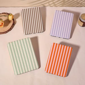 May include: Four rectangular cases with vertical striped patterns in brown, lavender, green, and orange. The cases are arranged on a light-colored surface. The cases appear to be for a tablet or e-reader.