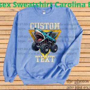 May include: Carolina blue unisex sweatshirt with a graphic of a shark-themed monster truck. The design includes the word "CUSTOM" above the truck and "TEXT" below. The sweatshirt is a solid color with a crew neck.