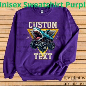 May include: Purple unisex sweatshirt featuring a shark-themed monster truck design. The graphic includes the words "CUSTOM TEXT" within a yellow triangle. The text "Unisex Sweatshirt Purple" is at the top. The bottom of the image says "the photos shown are just examples."