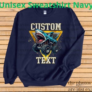 May include: Navy blue sweatshirt with a graphic of a shark-themed monster truck. The design includes the words "CUSTOM" and "TEXT" in a bold font, set against a yellow triangle. The text "Unisex Sweatshirt Navy" is at the top.