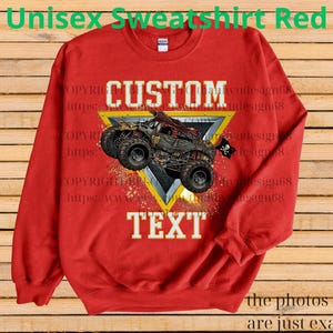 May include: A red unisex sweatshirt featuring a monster truck design. The graphic includes the word "CUSTOM" above the truck and "TEXT" below it, set against a yellow and silver triangle. The text "Unisex Sweatshirt Red" is at the top.