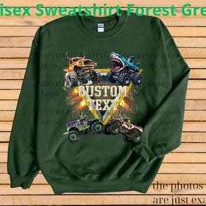 May include: Forest green unisex sweatshirt featuring a graphic of four monster trucks with the text "CUSTOM TEXT". The design includes a shark-themed truck and other trucks in orange, purple, and beige. The text "the photos shown are just examples" is also visible.