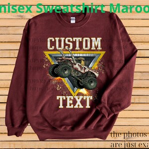May include: Maroon unisex sweatshirt with a custom design. The design features a monster truck graphic inside a yellow triangle, with the words "CUSTOM" and "TEXT". The text "the photos shown are just examples" is at the bottom.