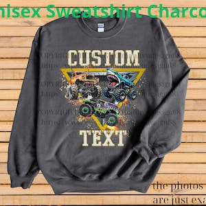 May include: Charcoal gray unisex sweatshirt featuring a graphic of three monster trucks in orange, blue, and green. The design includes the text "CUSTOM TEXT" within a yellow triangle. The sweatshirt has a crew neck.