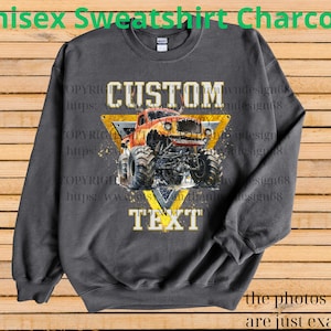May include: Charcoal gray unisex sweatshirt featuring a monster truck design. The design includes a yellow triangle with the words "CUSTOM" and "TEXT" and a monster truck illustration. The text "Unisex Sweatshirt Charcoal" is at the top.