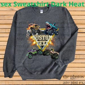 May include: Dark heather gray unisex sweatshirt featuring a graphic of monster trucks with the text "CUSTOM TEXT". The design includes a shark-themed truck and other colorful trucks. The text "Unisex Sweatshirt Dark Heather" is at the top.