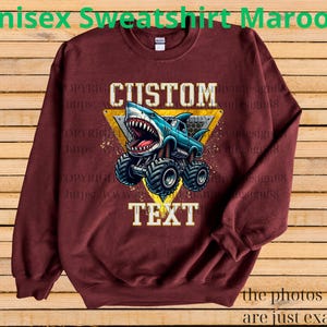 May include: Maroon unisex sweatshirt featuring a custom design. The design includes a blue monster truck with a shark head, set against a yellow triangle. The word "CUSTOM" is above the graphic, and "TEXT" is below.