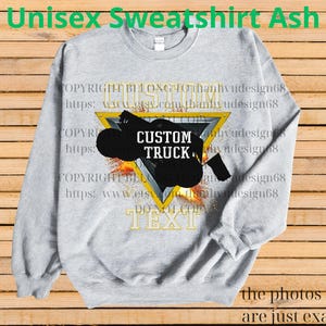 May include: Grey unisex sweatshirt with a black custom truck design inside a yellow triangle. The sweatshirt has the text "CUSTOM TRUCK" and other text. The background is a wooden surface.