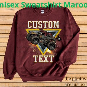 May include: Maroon unisex sweatshirt featuring a custom design. The design includes a monster truck graphic within a yellow triangle, with the words "CUSTOM" and "TEXT" in white. The sweatshirt has long sleeves and a crew neck.