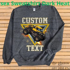 K&ouml;nnte beinhalten: Dunkelgraues Unisex-Sweatshirt mit einem gelb-schwarzen Monster-Truck-Motiv in einem gelben Dreieck. Die W&ouml;rter "CUSTOM" und "TEXT" sind &uuml;ber und unter der Grafik aufgedruckt. Der Text "the photos shown are just examples" steht unten.