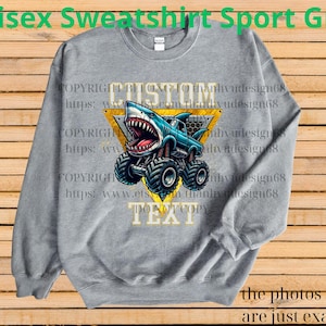 May include: A gray unisex sweatshirt with a shark monster truck graphic. The shark truck is blue and white with large black tires, set against a yellow triangle. The text "CUSTOM TEXT" is above and below the graphic. The words "Unisex Sweatshirt Sport Grey" are at the top.