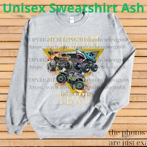 May include: Ash gray unisex sweatshirt featuring a graphic of three monster trucks in orange, blue, and green. The design includes a yellow triangle with the text "CUSTOM TEXT" and copyright information. The sweatshirt has long sleeves and a crew neck.