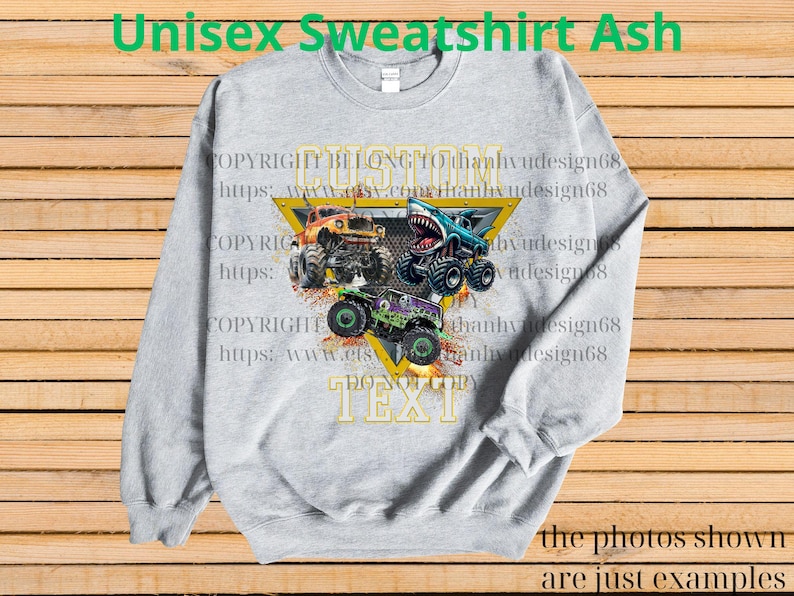 May include: Ash gray unisex sweatshirt featuring a graphic of three monster trucks in orange, blue, and green. The design includes a yellow triangle with the text "CUSTOM TEXT" and copyright information. The sweatshirt has long sleeves and a crew neck.