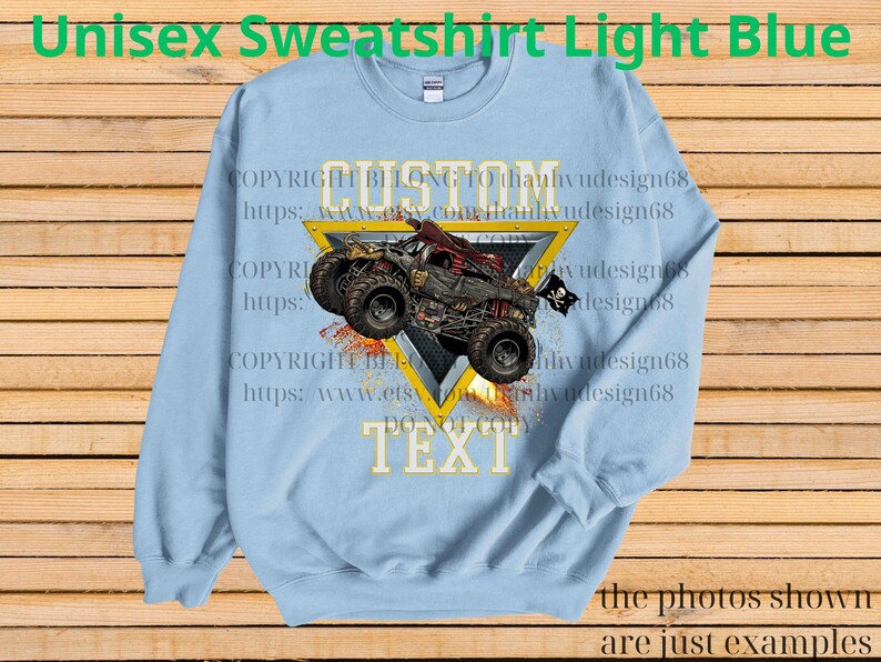 May include: Light blue unisex sweatshirt with a monster truck graphic. The design includes a red and black monster truck with a skull and crossbones flag, set against a yellow triangle. The text "CUSTOM TEXT" is also visible.