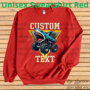 May include: Red unisex sweatshirt featuring a shark-themed monster truck design. The graphic includes the words "CUSTOM TEXT" in white, set against a yellow triangle. The sweatshirt has long sleeves and a crew neck.