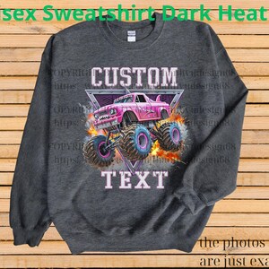 Op de afbeelding: Donkergrijs unisex sweatshirt met een roze monster truck afbeelding. De afbeelding bevat de woorden "CUSTOM" en "TEXT" in een lichtroze lettertype, met een driehoekige achtergrond.
