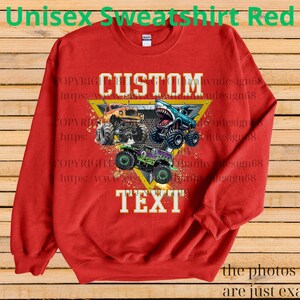 May include: Red unisex sweatshirt with a graphic of three monster trucks. The sweatshirt has the words "CUSTOM TEXT" in white block letters. The top of the sweatshirt has the words "Unisex Sweatshirt Red" in green.